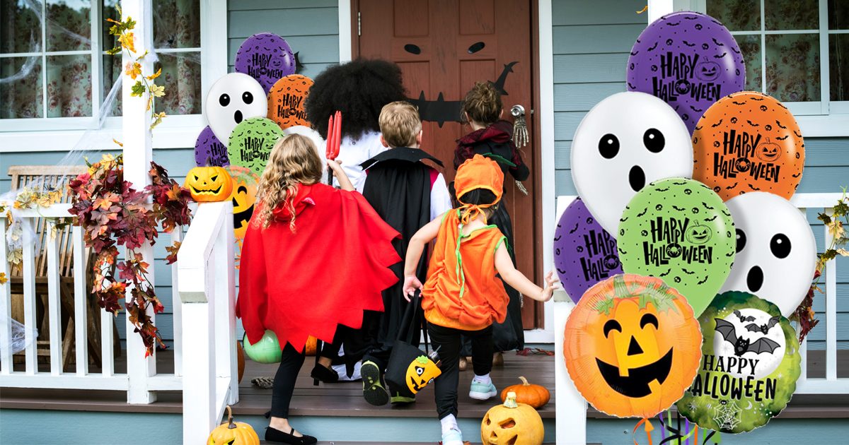 Kids in costumes trick-or-treat at a decorated house with pumpkins and Halloween balloons featuring ghosts and pumpkins.