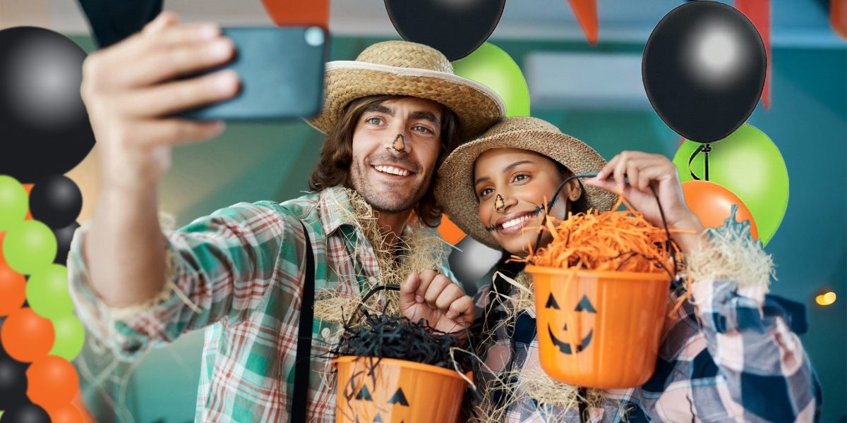 Couple dressed as scarecrows taking a selfie at a Halloween party with pumpkin buckets and festive black, orange, and lime green balloon decorations