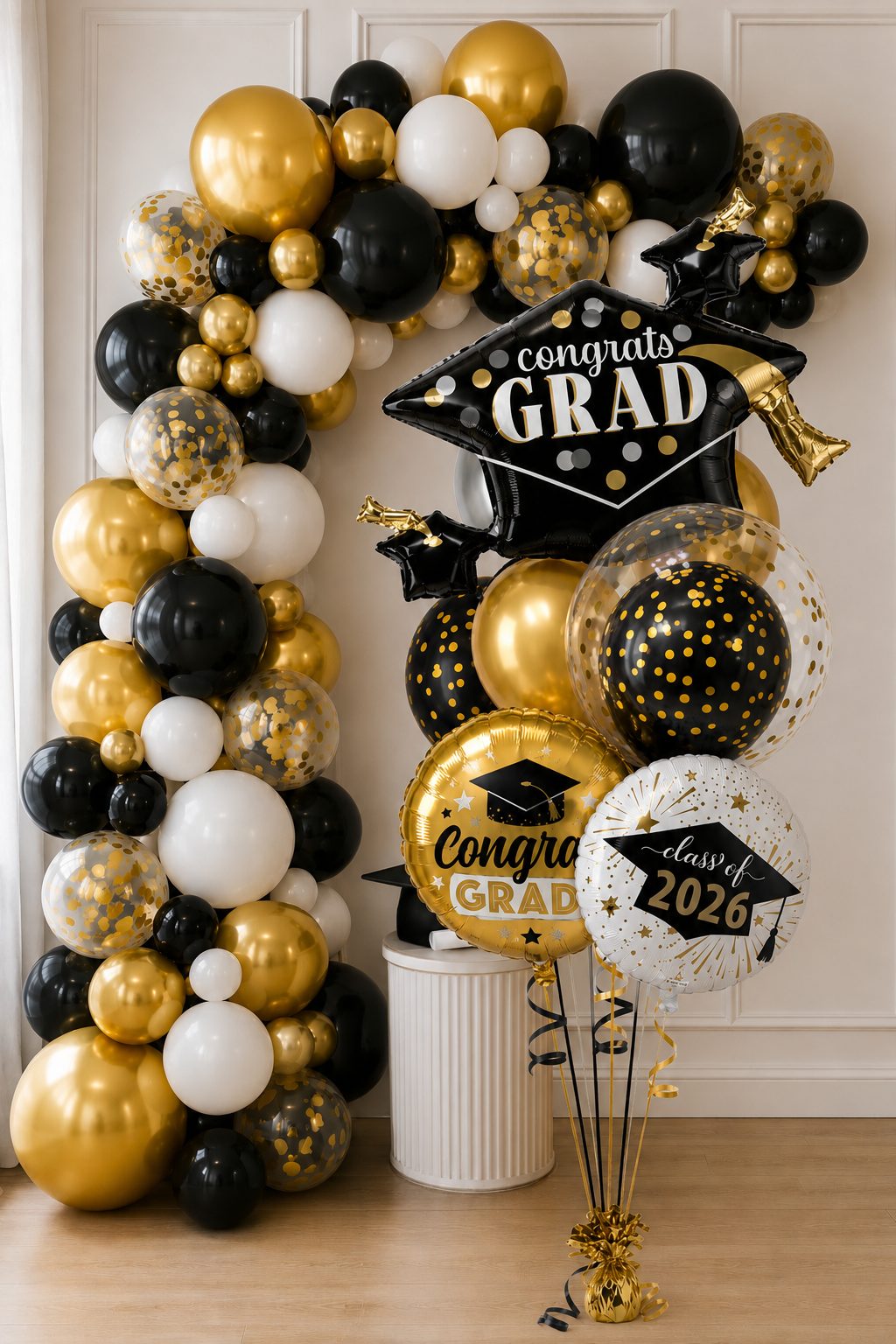 A tall, dramatic graduation balloon bouquet in a black, gold, and white color scheme, featuring a large black "Congrats Grad" graduation cap balloon and a round "Congratulations Graduate" balloon, styled indoors against a white paneled wall.