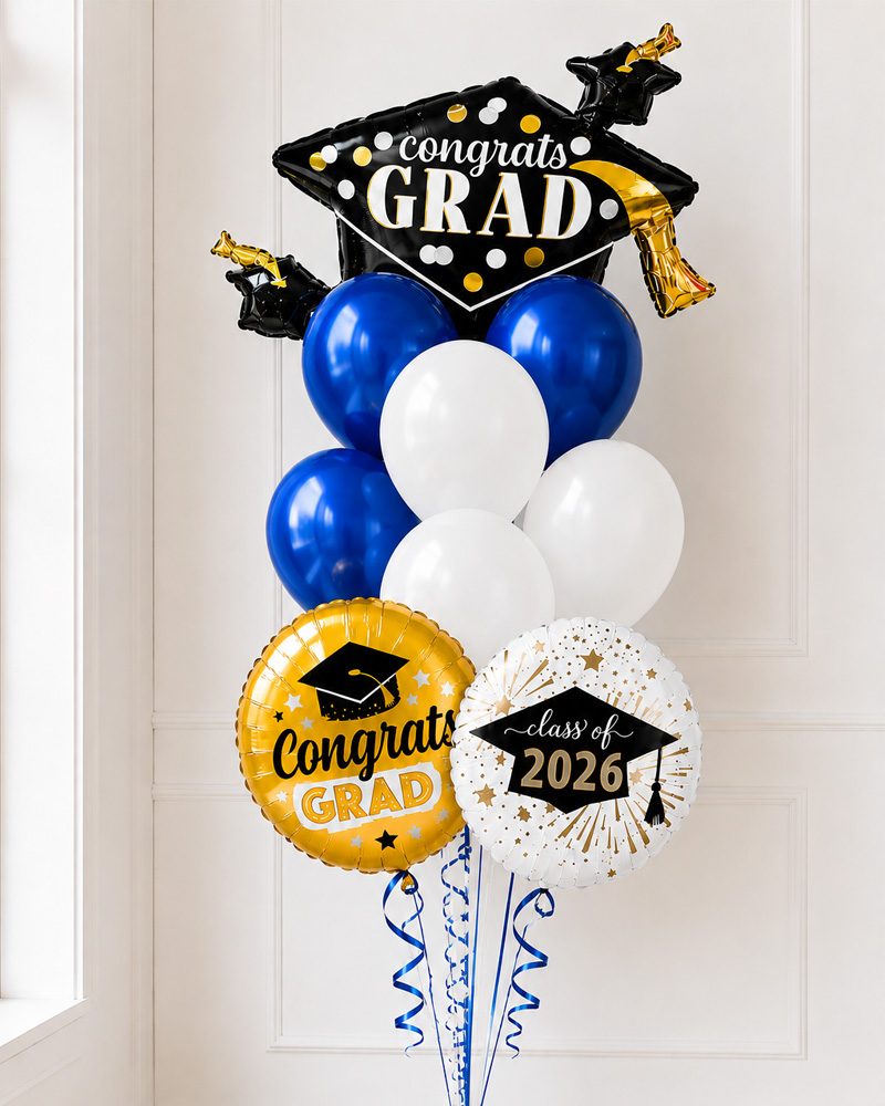 Graduation balloon bouquet in navy blue, white, and gold displayed against a clean white paneled wall with soft natural lighting. Features a large black “Congrats Grad” graduation cap foil balloon, blue and white latex balloons, gold and white graduation-themed mylar balloons, curling blue ribbons, and a metallic blue foil weight at the base. Portrait orientation, photorealistic style, bright and airy indoor setting with a professional product photography aesthetic.